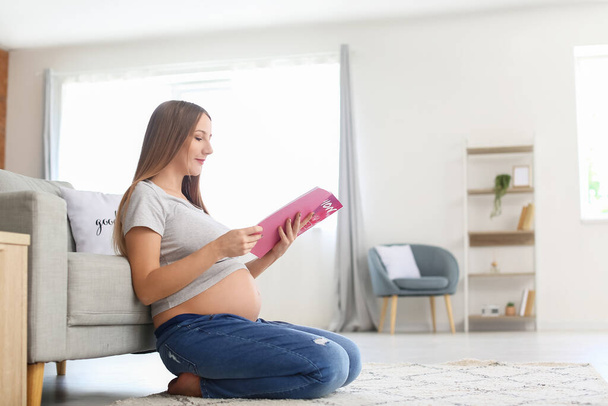Νεαρή έγκυος γυναίκα διαβάζει περιοδικό μόδας στο σπίτι - Φωτογραφία, εικόνα