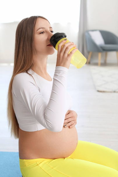 Junge schwangere Frau in stylischer Sportkleidung trinkt zu Hause Wasser - Foto, Bild