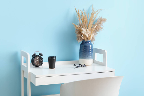 Table with dried spikelets in vase, alarm clock, cup of coffee and eyeglasses near blue wall - Foto, Bild