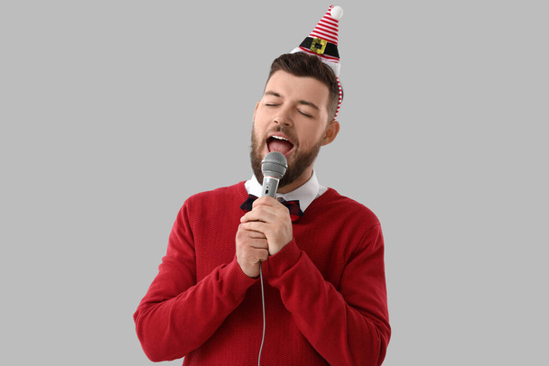 Young man in Santa hat singing with microphone on grey background - Photo, Image