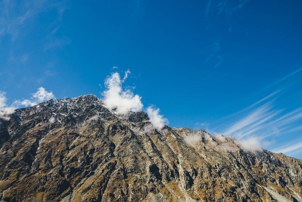 Scenic herfst zonnig landschap met hoge rotsachtige berg met piektop in lage wolken onder de blauwe hemel. Geweldige alpine uitzicht op mooie scherpe top in de zon. Prachtige bergtop in zonlicht. - Foto, afbeelding