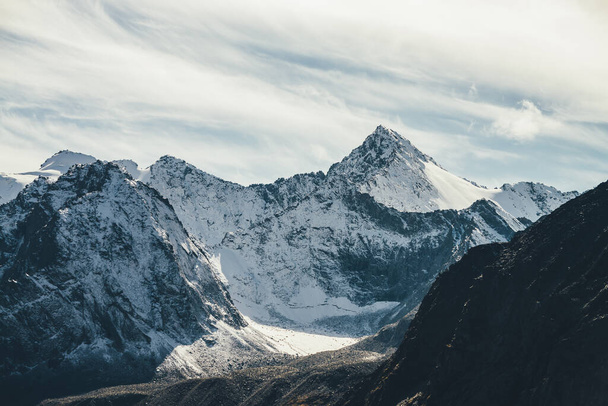 Toller Blick auf die hoch verschneite Bergwand mit Gipfel unter Zirruswolken am Himmel. Alpine Landschaft mit großen schneebedeckten Bergen mit scharfem Gipfel in der Sonne. Weiß-schneebedeckter Gipfel im Sonnenlicht. - Foto, Bild