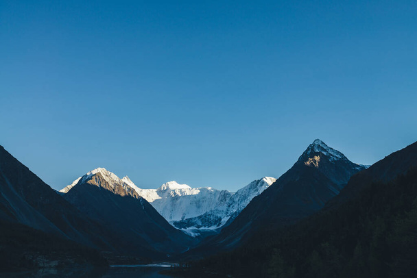Tolle alpine Landschaft mit Bergsilhouetten und schneebedeckten Berggipfeln in goldenem Sonnenschein unter klarem blauen Himmel. Schöne sonnenbeschienene schneeweiße Bergwand und See im schattigen Hochlandtal - Foto, Bild