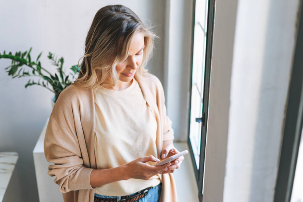 Молодая улыбающаяся блондинка с длинными волосами в стильном кардигане работает за ноутбуком в ярком современном офисе, женщина отправляет сообщение на смартфоне - Фото, изображение