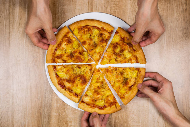 Top view of hands taking slices of delicious and crispy hawaiian pizza topping with mozzarella cheese, pineapple and tomato source. Group of hungry friends sitting at desk and sharing delicious lunch on wooden texture table background.  - Fotó, kép