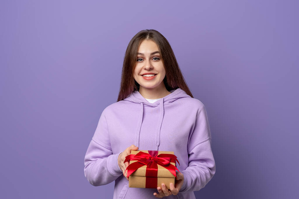 Giovane donna bruna felice sorriso tenere regalo scatola decorata con nastro rosso in mano, in piedi su sfondo viola. Il concetto di festa di compleanno di Capodanno della donna - Foto, immagini