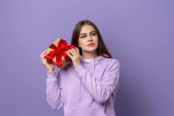 Glückliche junge brünette Frau in lässigem Kapuzenpulli, die isoliert vor lila Hintergrund steht und eine Geschenkschachtel in der Hand hält. Neujahrsempfang zum Frauentag - Foto, Bild