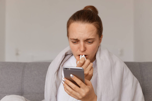 Indoor shot of upset ill woman with hair bun sitting on sofa wrapped in blanket having influenza symptoms, using smart phone, reading about treatment in internet, - Foto, Imagem