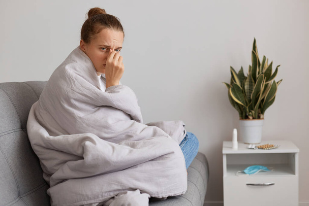 Sick sad woman with hair bun sitting on sofa wrapped in blanket fells unwell, posing at home, has tired and upset expression, suffering influenza, grippe or fever. - Photo, Image