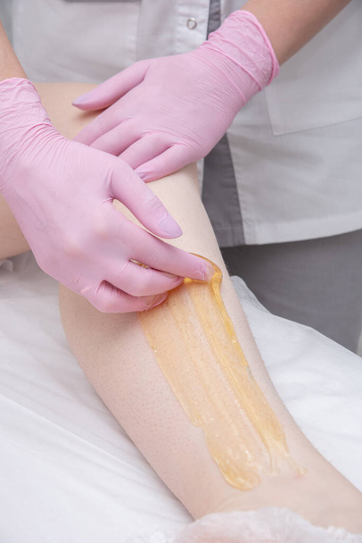 Proces depilacji cukru. Mistrz w różowych rękawiczkach medycznych nakłada żółtą pastę na nogę klienta. - Zdjęcie, obraz