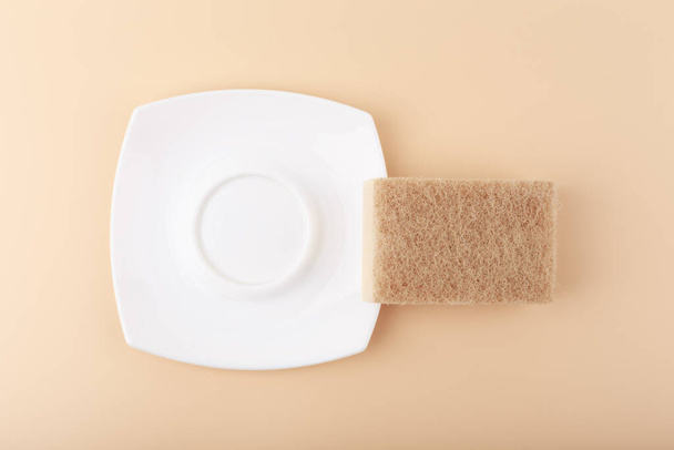 Dishwashing minimal creative concept in light beige colors. White ceramic plate and cleaning sponge top view - Fotoğraf, Görsel