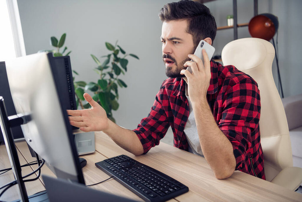 Zdjęcie nieszczęśliwego młodego człowieka trzymać telefon rozmawiać redaktor wygląd ekran siedzieć stół wewnątrz biura w miejscu pracy - Zdjęcie, obraz