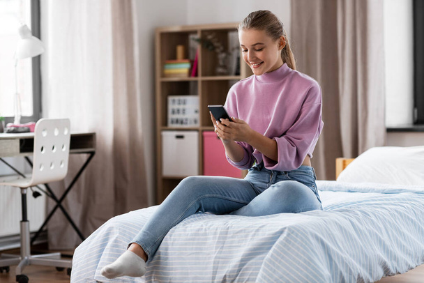 szczęśliwa dziewczyna ze smartfonem siedzi na łóżku w domu - Zdjęcie, obraz