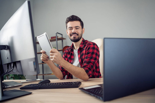 Photo of happy positive young man hold tablet sit desk pc computer it specialist indoors inside office - Foto, imagen