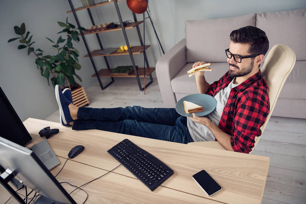 Portrait of attractive trendy cheery guy eating snack watching monitoring security camera legs on table rest at office work place station indoor - Foto, Imagen