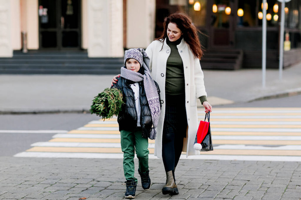 Μια όμορφη μητέρα και ο γιος της βγαίνουν από το μαγαζί, κρατώντας μια τσάντα με δώρα και ένα μικρό χριστουγεννιάτικο δέντρο. Ψώνια για τις διακοπές. Η έννοια των Χριστουγέννων και της Πρωτοχρονιάς - Φωτογραφία, εικόνα