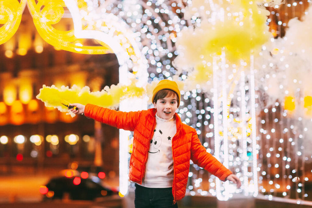 Nastoletni chłopiec z bombą dymną w rękach podczas świąt Bożego Narodzenia lub Nowego Roku na tle miejskiego oświetlenia - Zdjęcie, obraz