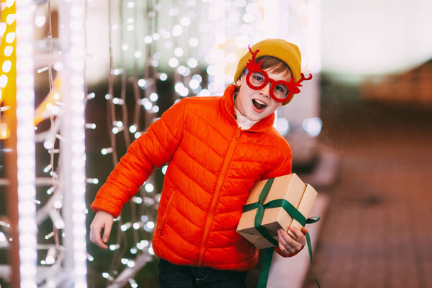 Un niño alegre con una chaqueta naranja y gafas divertidas camina por la ciudad de la noche con iluminación festiva. Regalos de Navidad, preparación para las fiestas - Foto, imagen