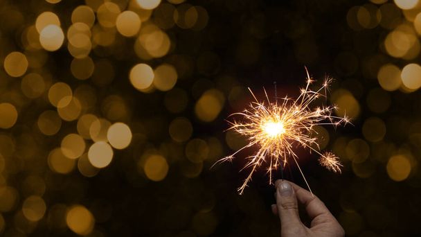 Silvester Party / New Year celebration festive background banner - Young happy pretty woman holding a sparkling sparkler in her hand at dark night with golden bokeh - Photo, Image