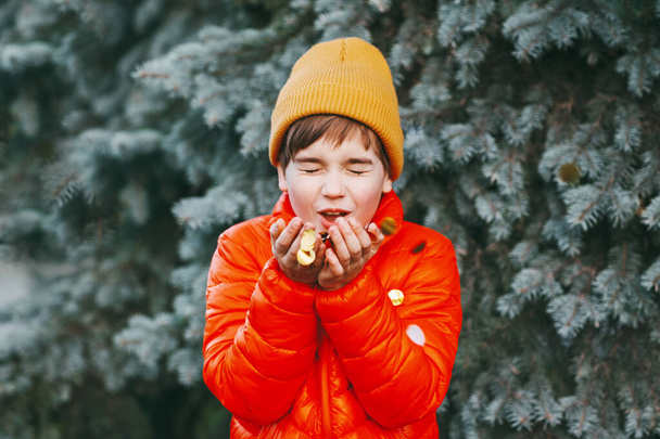 Ein Junge in orangefarbener Jacke und Mütze hält glänzendes Konfetti in den Händen und pustet drauf, der Junge freut sich auf die Feiertage. Feiertagsstimmung, Weihnachten, Neujahr - Foto, Bild