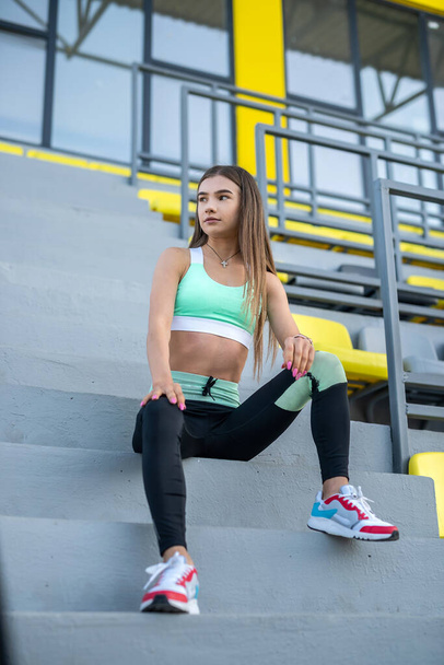 Молодая женщина в спортивной форме сидит на стуле стадиона и резр после утренних упражнений - Фото, изображение