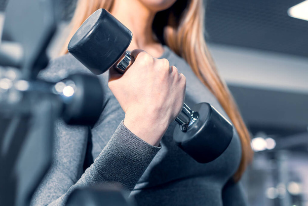 girl lifts a dumbbell, the process of training a girl in a gray suit for fitness. the background is blurry, out of focus - Photo, Image
