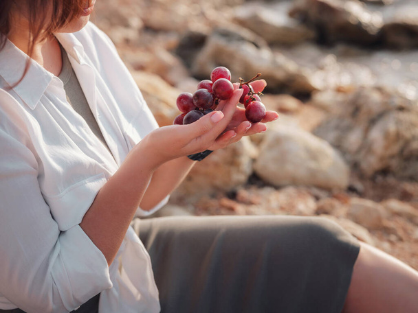 Тропический пикник у моря. Счастливая молодая женщина наслаждается пикником с видом на океан, сидеть на скале пить розовое вино. пикник с вином, сыром и свежим виноградом на пляже на закате. - Фото, изображение