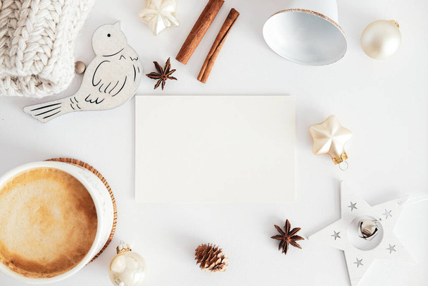 Hygge Christmas composition. Flat lay blank greeting card mockup, cinnamon sticks, cup of coffee, bell, balls, knitted hat, wooden Christmas decorations on white table. Top view. Nordic style. - Foto, Imagen