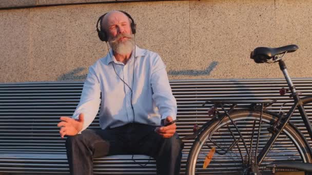Funny gris pelo bigote barbudo ciclista se sienta en el banco de la ciudad descansando después del viaje. Anciano activo en edad de jubilación, con auriculares, escuchando música alegre, cantando disfrutando de un fin de semana soleado - Metraje, vídeo
