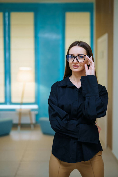 Attractive woman in dark shirt and glasses standing arms crossed in hall and smiling. Female appearance concept. Full length view - Fotoğraf, Görsel