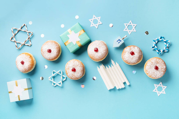 Hanukkah sweet doughnuts sufganiyot (traditional donuts) with fruit jelly jam, gift boxes, spinnig driedel and candles on blue paper background. Jewish holiday Hanukkah concept. Top view, copy space. - Photo, Image