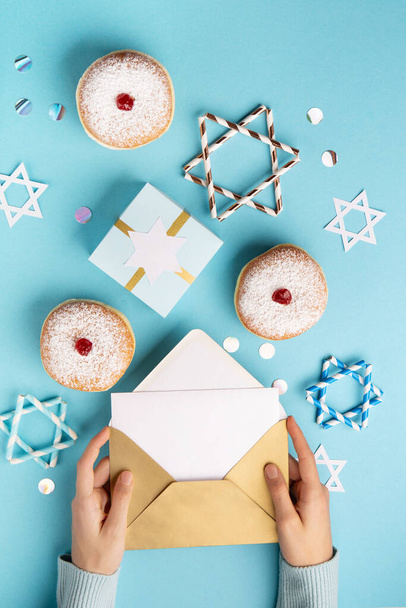 Hanukkah sweet doughnut sufganiyot with powdered sugar and fruit jam and empty white letter in woman's hands on blue paper background. Jewish holiday Hanukkah concept. Top view, copy space - Foto, Imagem