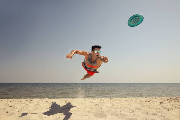Sportive man jumping while trying to catch flying disk at beach - Photo, Image
