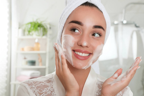 Красивая молодая женщина наносит пену на лицо в ванной комнате. Косметический уход за кожей - Фото, изображение