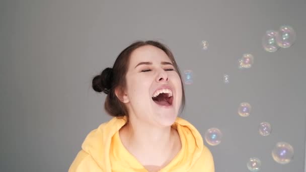 happy brunette woman in yellow hoodie among soap bubbles in bright grey room. happy people. millennial generation. fashionable teenager. trendy colours. slow motion. - 映像、動画