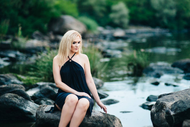 blond meisje in een zwart jurk met blauwe ogen op de stenen kust van de rivier - Foto, afbeelding