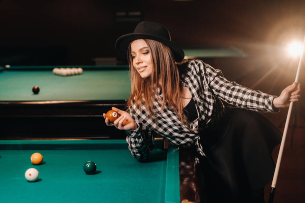 A girl in a hat in a billiard club with a cue and balls in her hands.Playing pool. - Photo, Image