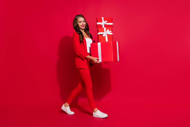 Full size profiel foto van schattig brunette dame hold cadeautjes gaan dragen pak sneakers geïsoleerd op rood kleur achtergrond. - Foto, afbeelding