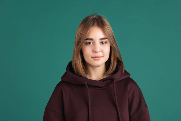 Half-length portrait of young charming girl, student in brown hoodie looking at camera isolated on green vintage studio backgroud. - Фото, зображення