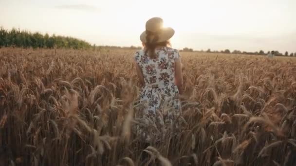 Une agréable jeune femme en chapeau et robe d'été blanche traverse un champ de blé et se tourne vers la caméra en souriant. Mouvement lent - Séquence, vidéo