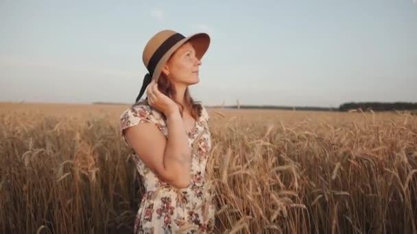 A tender young woman stands in a wheat field and looks to the sky putting your hand over the hat. The concept of freedom and calmness - Footage, Video