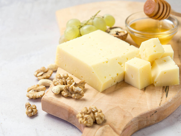 queso de calidad agrícola se encuentra en una tabla de madera rodeada de uvas. nueces y miel primer plano sobre un fondo claro - Foto, Imagen