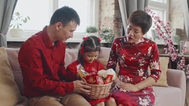 Colpo medio di felice padre e madre asiatici in camicia rossa e abito tradizionale dando cinese nuovo anno cesto regalo per la loro carina figlia di 5 anni mentre celebrano insieme a casa - Filmati, video