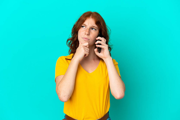 Teenager reddish woman using mobile phone isolated on blue background having doubts and thinking - Fotografie, Obrázek