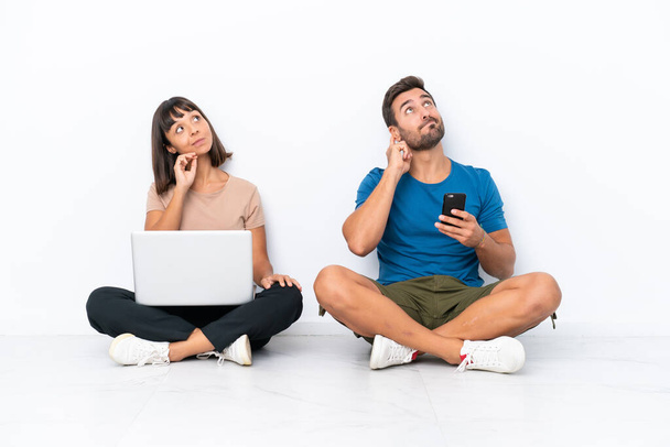 Νεαρό ζευγάρι κάθεται στο πάτωμα κρατώντας το PC και το κινητό τηλέφωνο απομονώνονται σε λευκό φόντο σκέφτεται μια ιδέα, ενώ ξύσιμο κεφάλι - Φωτογραφία, εικόνα