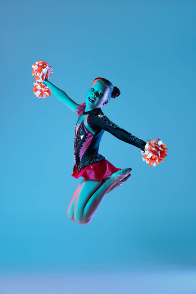 Full-length portret van mooi meisje, cheerleader training, optreden in helder bodysuit met pompons geïsoleerd over blauwe achtergrond in neon lichten - Foto, afbeelding