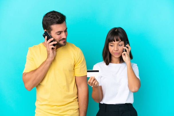 Jeune couple isolé sur fond bleu achetant avec le mobile avec une carte de crédit - Photo, image