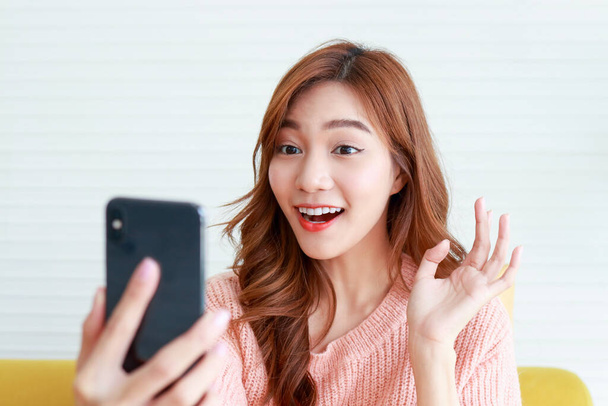 piękna azjatycka kobieta uśmiechnięta szczęśliwa na żywo w domu Trzymając czarny smartfon, online rozmowy wideo, rozmowy z przyjaciółmi przez Internet. Nowoczesna koncepcja technologii komunikacyjnych, odległość społeczna - Zdjęcie, obraz