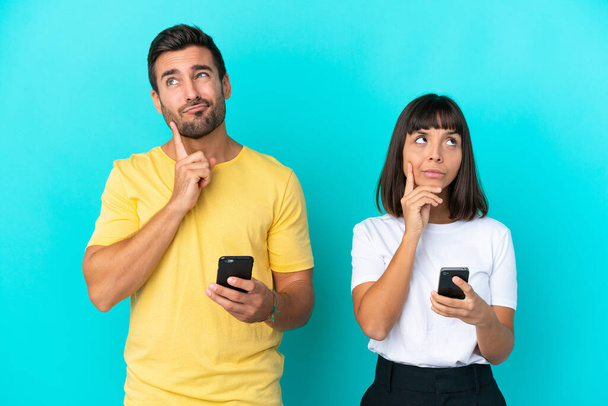 Молодая пара изолированы на синем фоне с помощью мобильного телефона и думать - Фото, изображение