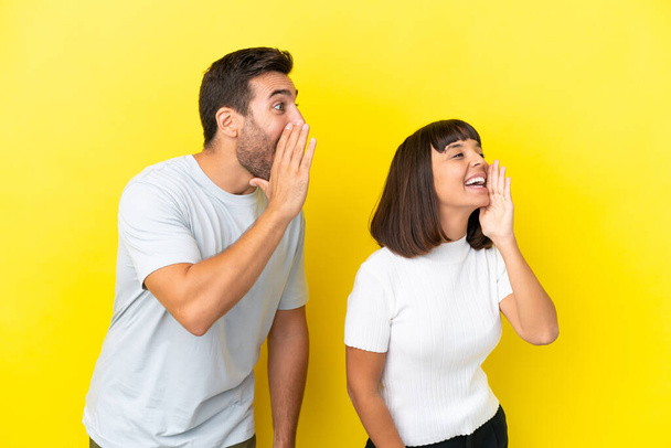 Young couple isolated on yellow background shouting with mouth wide open to the lateral - 写真・画像